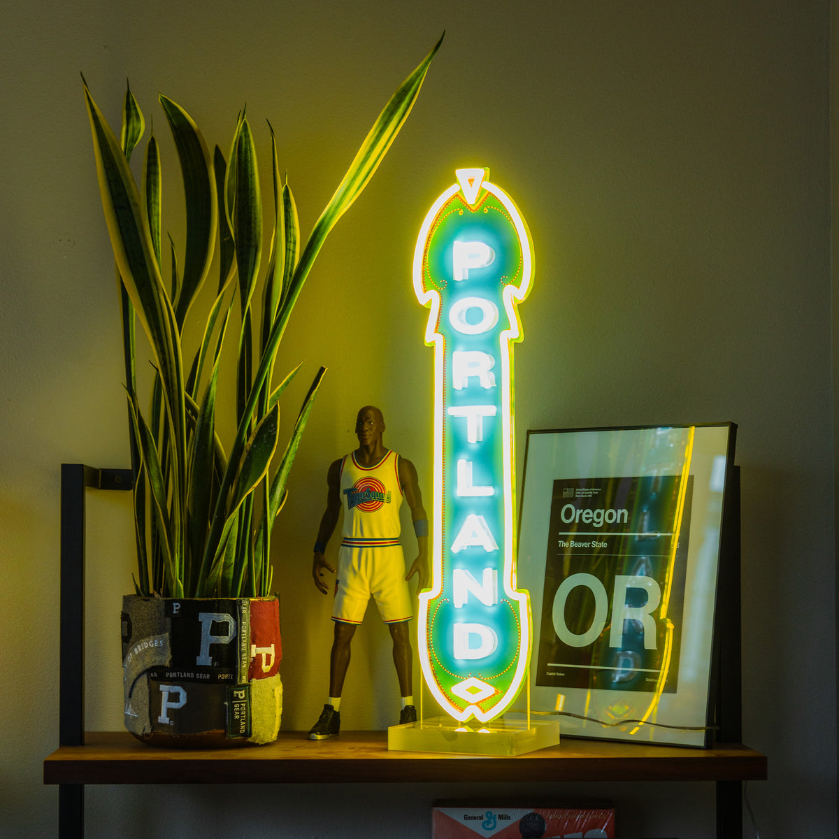 A shelf featuring a potted plant, a basketball player figurine, an energy-efficient yellow LED Marquee Sign that reads "Portland," and a framed poster of Oregon creates a vibrant, eclectic display. The neon sign and the plant work together to stand out against the dimly lit background wall, highlighting the bright elements on the shelf.