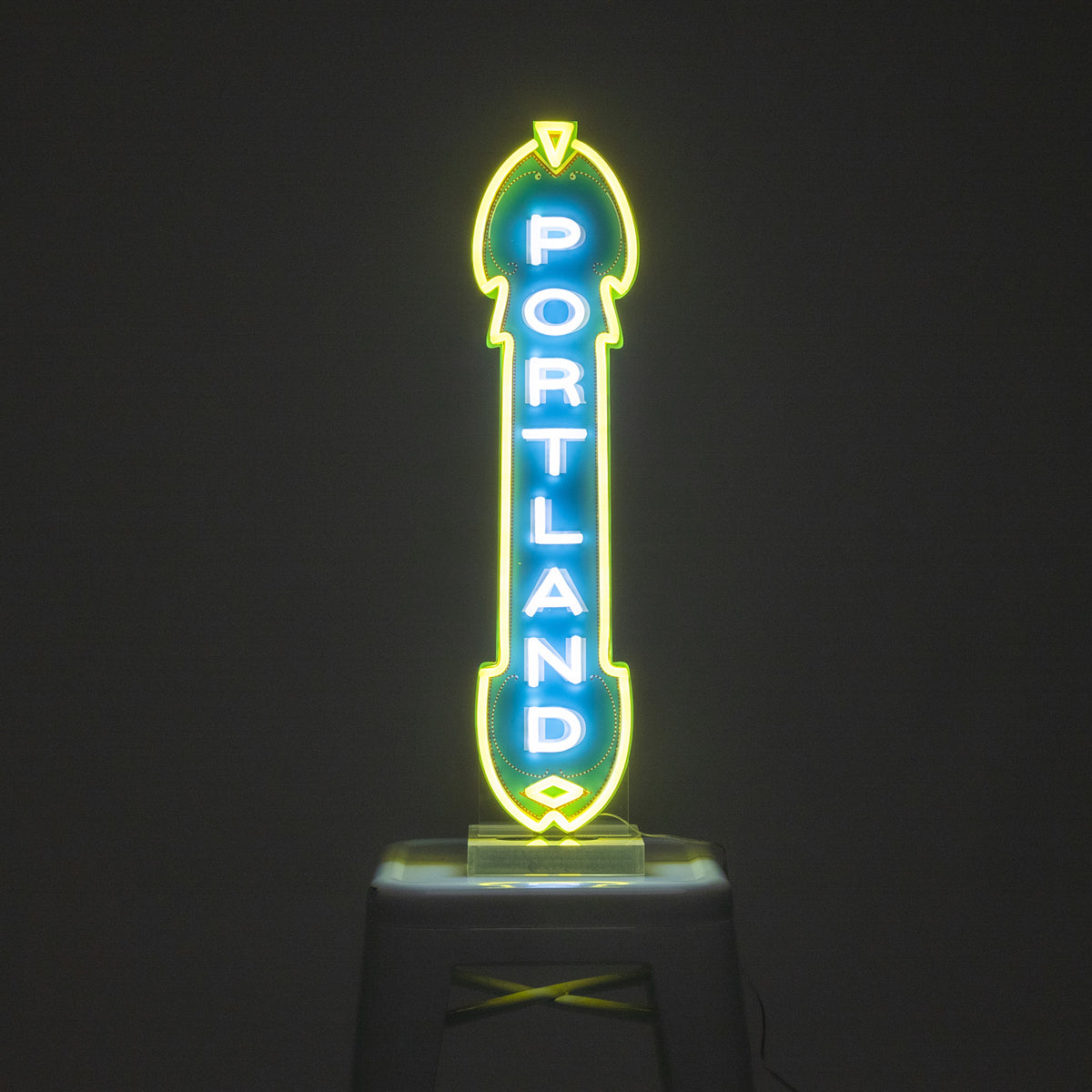 A custom LED Marquee Sign in the shape of an arrow, glowing in green, blue, and yellow, with the word "PORTLAND" written vertically in the center. This energy-efficient sign is displayed against a dark background and placed on a stool.